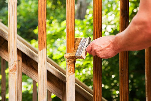A male hand, holding a large paintbrush, is applying stain to a weathered and cleaned treated pine deck railing. The background is sunlit trees and bushes.